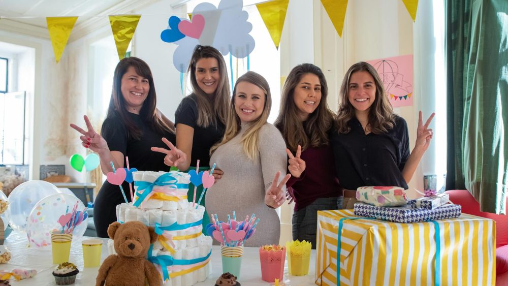 Smiling friends celebrate a baby shower London, surrounded by gifts, decorations, and a diaper cake in a bright, festive setting.